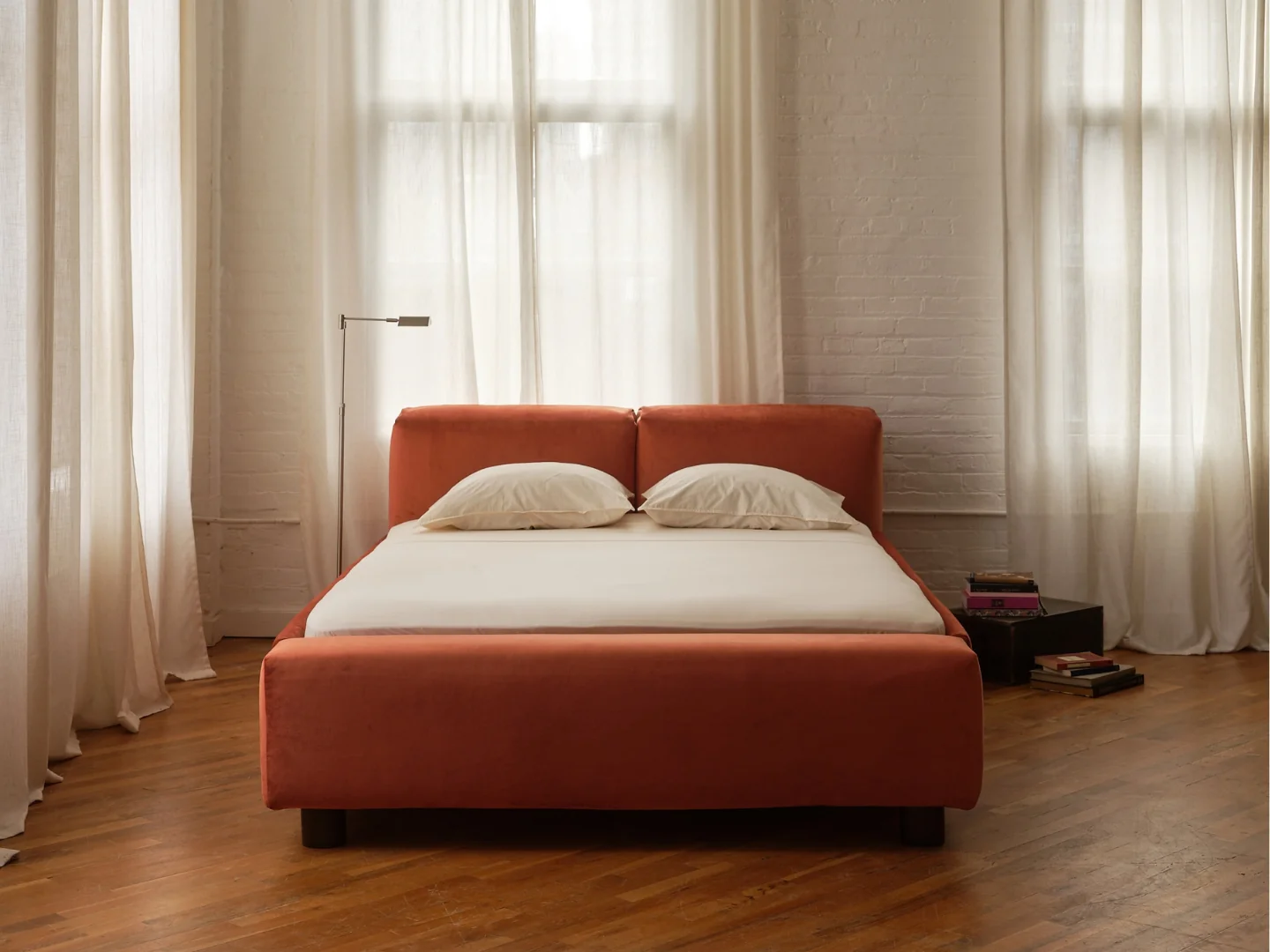 Low-profile upholstered bed frame in burnt orange with white bedding, positioned against a white brick wall between two windows.