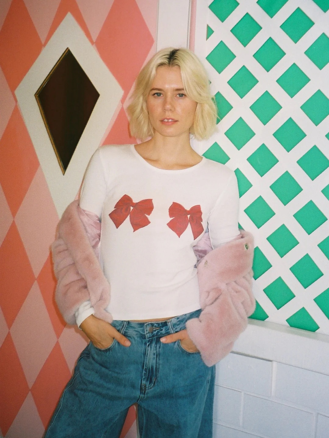 A person with short blonde hair wears a white shirt with red bows, blue jeans, and a light pink jacket, standing against a geometric pastel wall.
