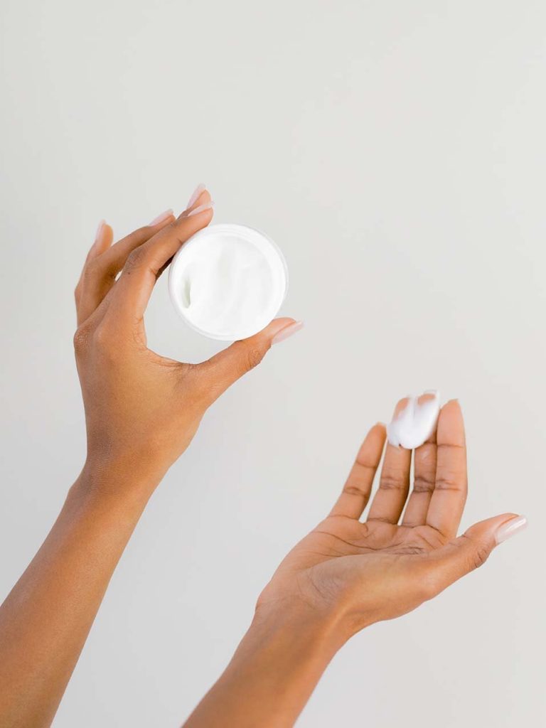 A person holds a container of white cream in one hand and applies some to the back of their other hand against a plain background.