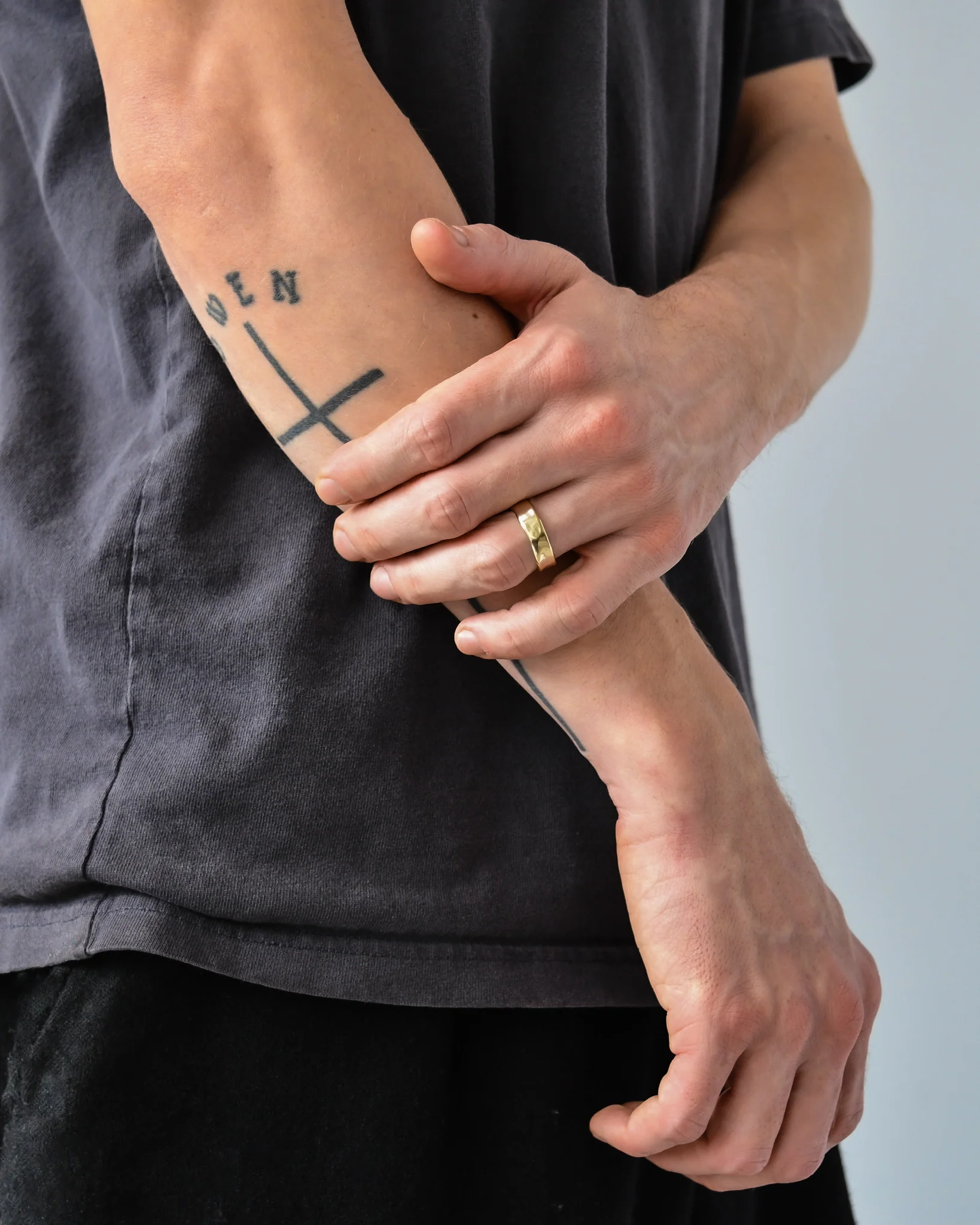 Person wearing a gold ring on their left hand holds their right forearm, which has a tattoo of a cross and the letters “DEN.”.