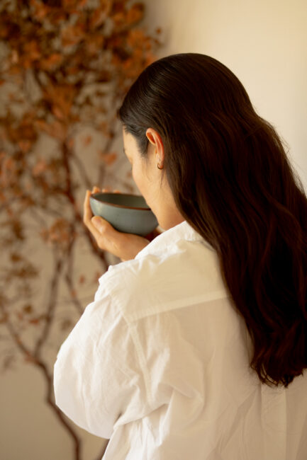 A person with long dark hair, wearing a white shirt, holds a bowl close to their face, standing near a wall with a dried branch decoration.