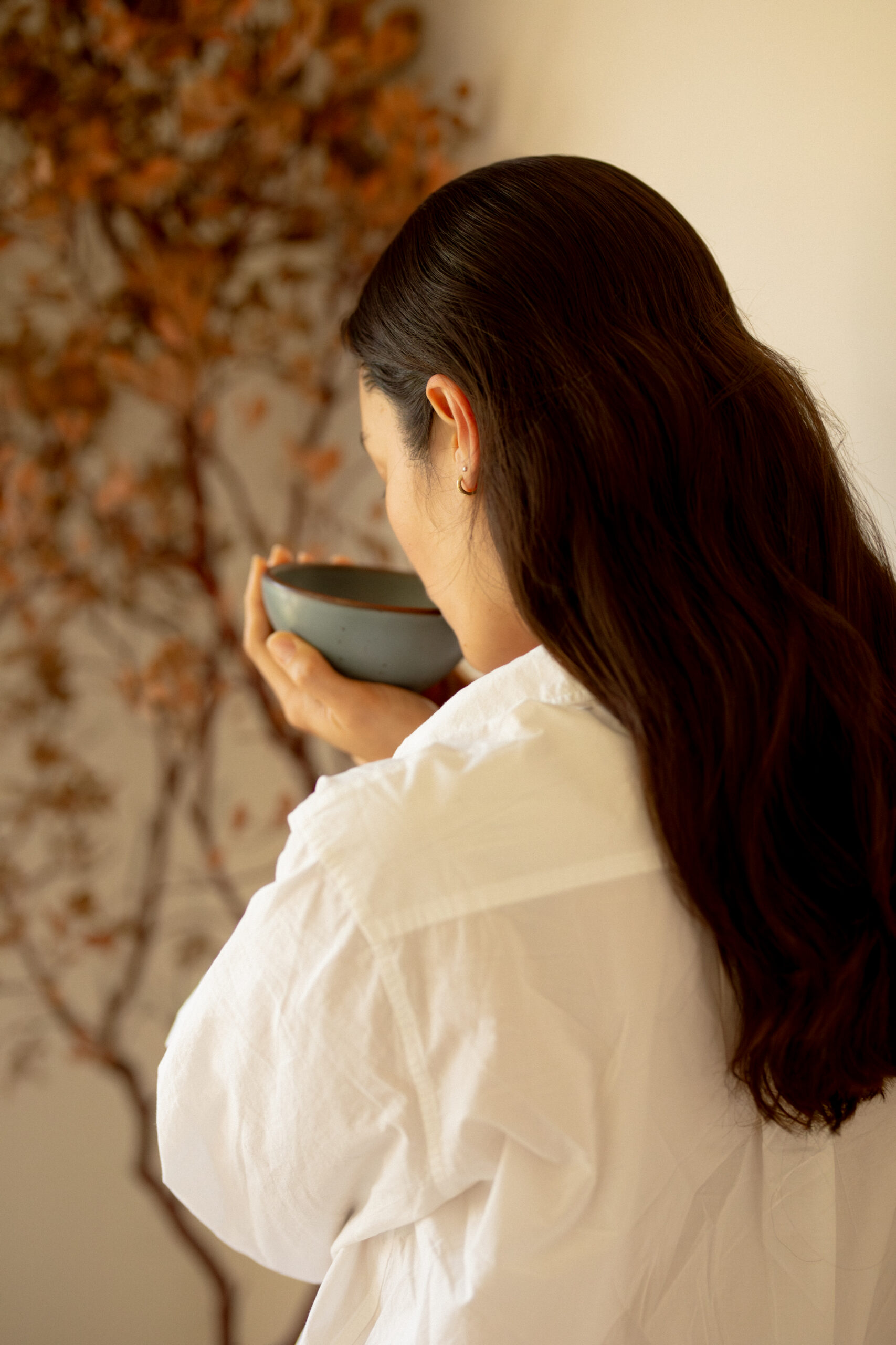 A person with long dark hair, wearing a white shirt, holds a bowl close to their face, standing near a wall with a dried branch decoration.