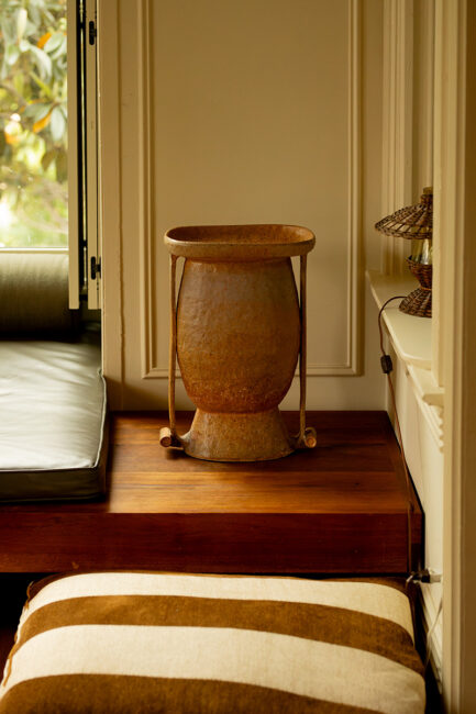 A rusty metal vase sits on a wooden platform beside a window, with a striped cushion and a small woven lamp nearby.