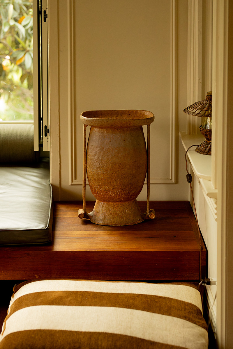 A rusty metal vase sits on a wooden platform beside a window, with a striped cushion and a small woven lamp nearby.