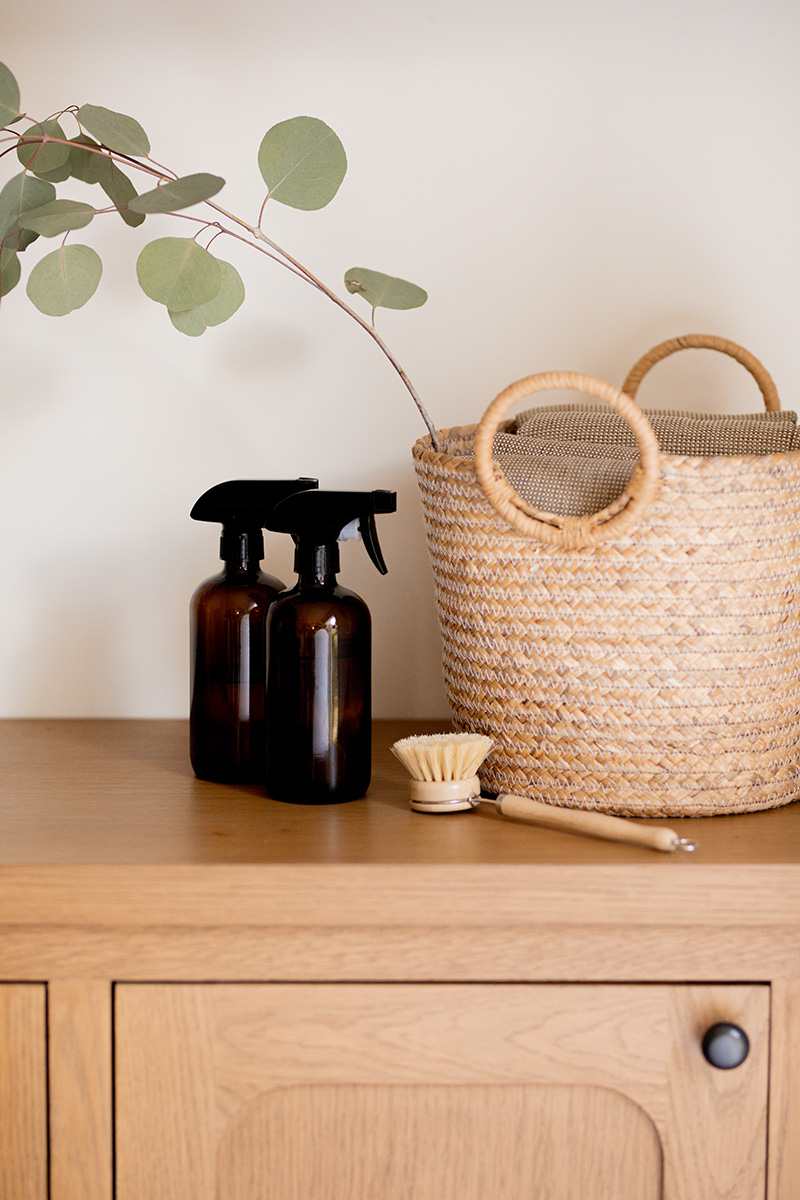 Two brown spray bottles, a small cleaning brush, and a woven basket with handles sit on a wooden cabinet next to a branch with green leaves.