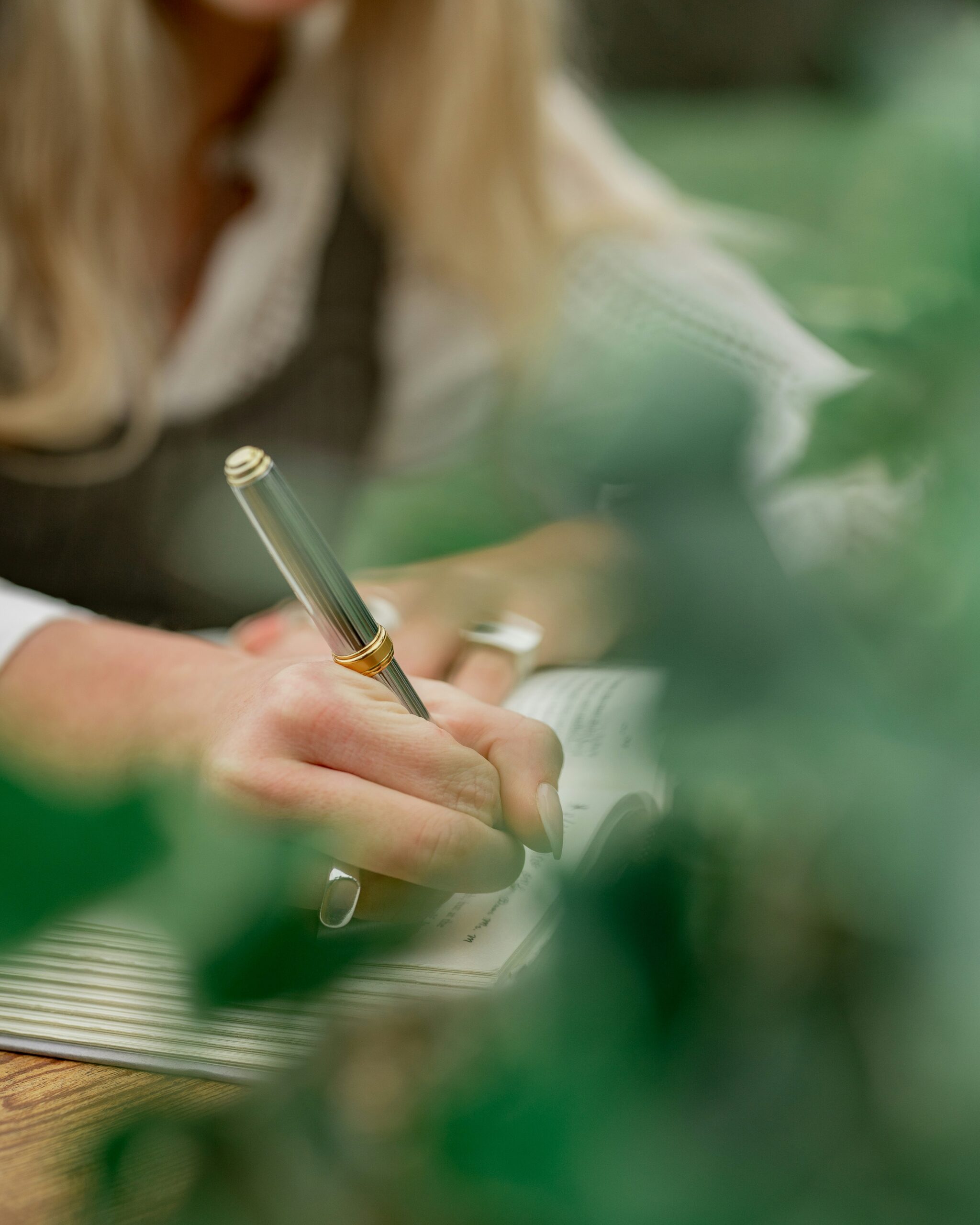 A person with long hair writes in a notebook with a silver pen, partially obscured by green leaves in the foreground.