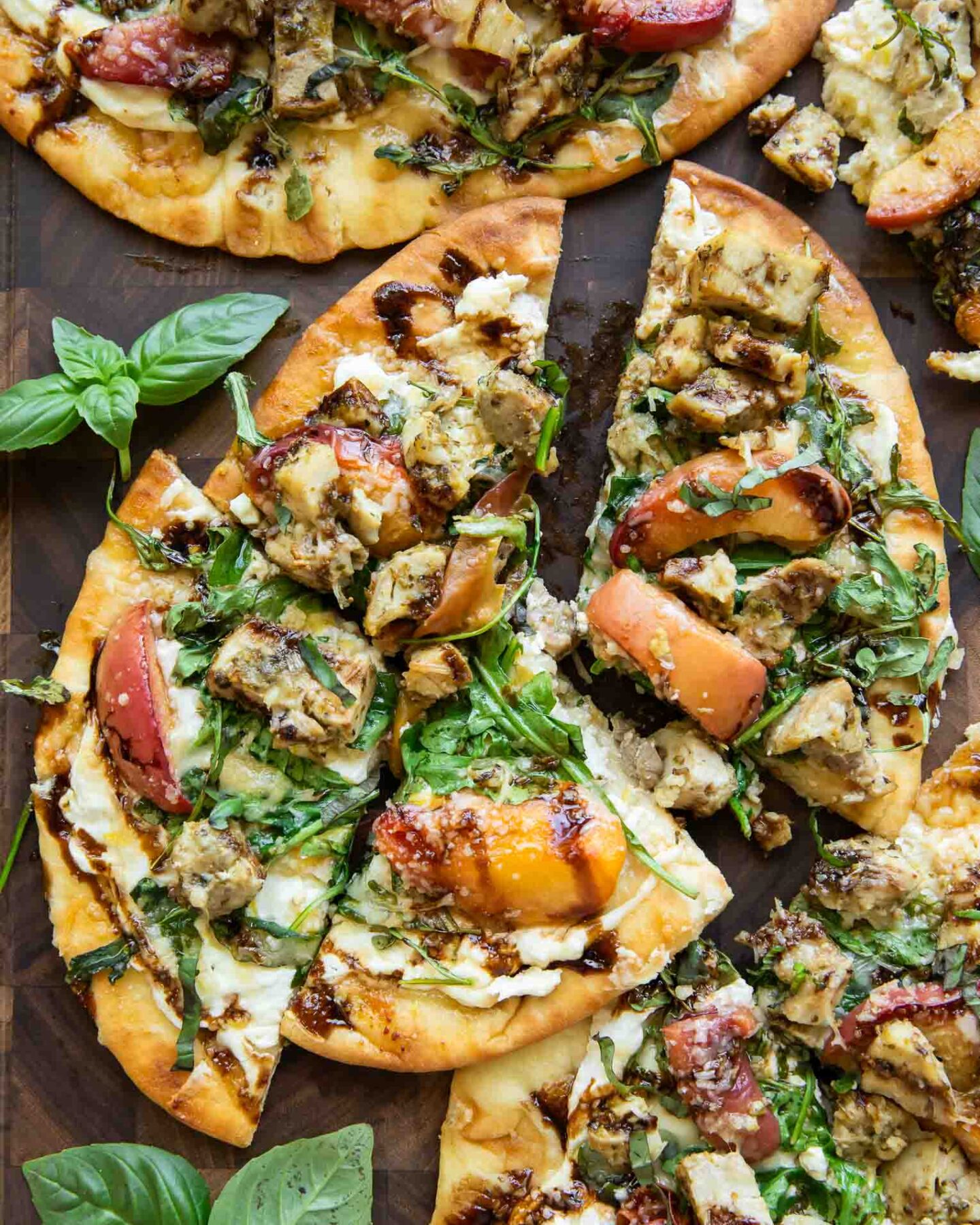 Flatbread pizzas topped with grilled chicken, arugula, sliced peaches, cheese, and a drizzle of balsamic glaze, arranged on a wooden surface with fresh basil leaves nearby.