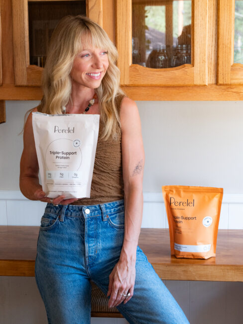 Woman holding a bag of Perelel Triple-Support Protein, standing by a wooden counter with another protein bag placed beside her.