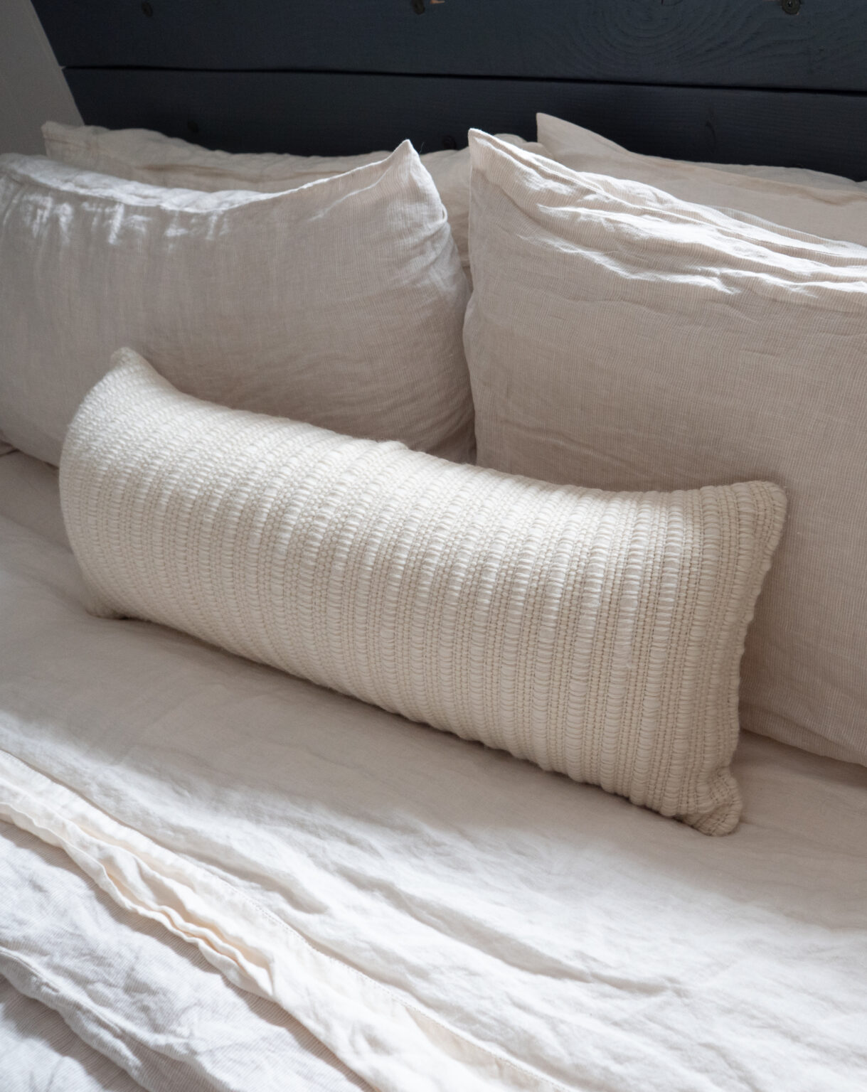 A long, textured white pillow rests on a neatly made bed with two large white pillows and white bedding.