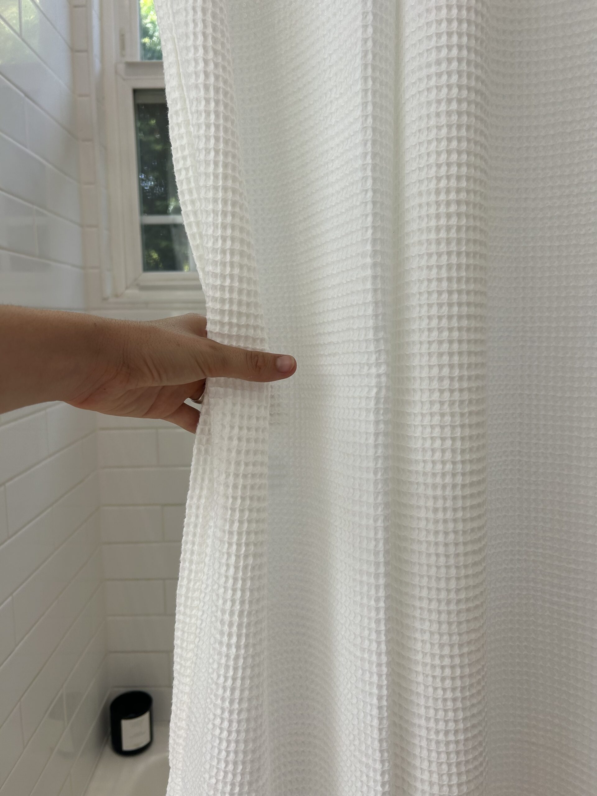 A hand pulls back a textured white shower curtain in a tiled bathroom with a small black container on the floor.
