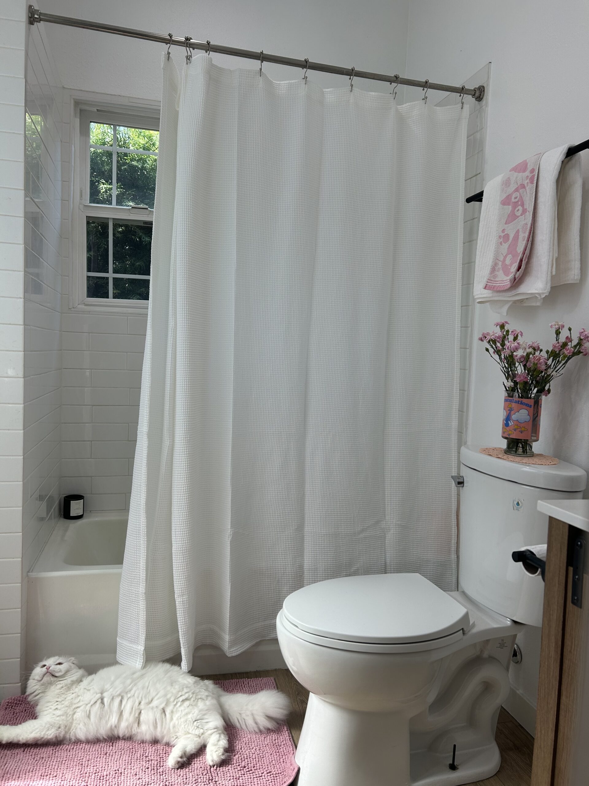 A white cat lies on a pink rug next to a toilet in a bathroom with a white shower curtain, flowers on the toilet tank, and towels on a rack.