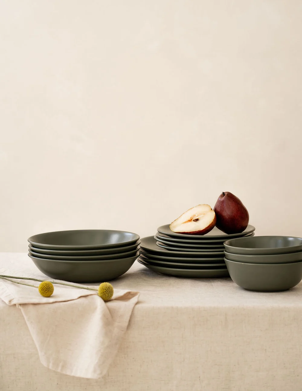 Stacked green ceramic bowls and plates on a beige tablecloth, with a halved pear and small yellow flowers arranged nearby.