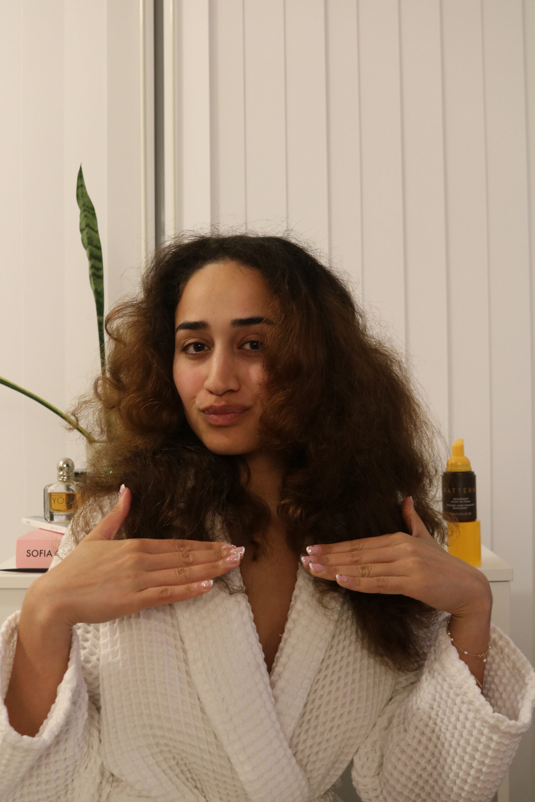 A person with long, curly hair wears a white robe, showing their manicured nails. Skincare and hair products are visible on a shelf in the background.