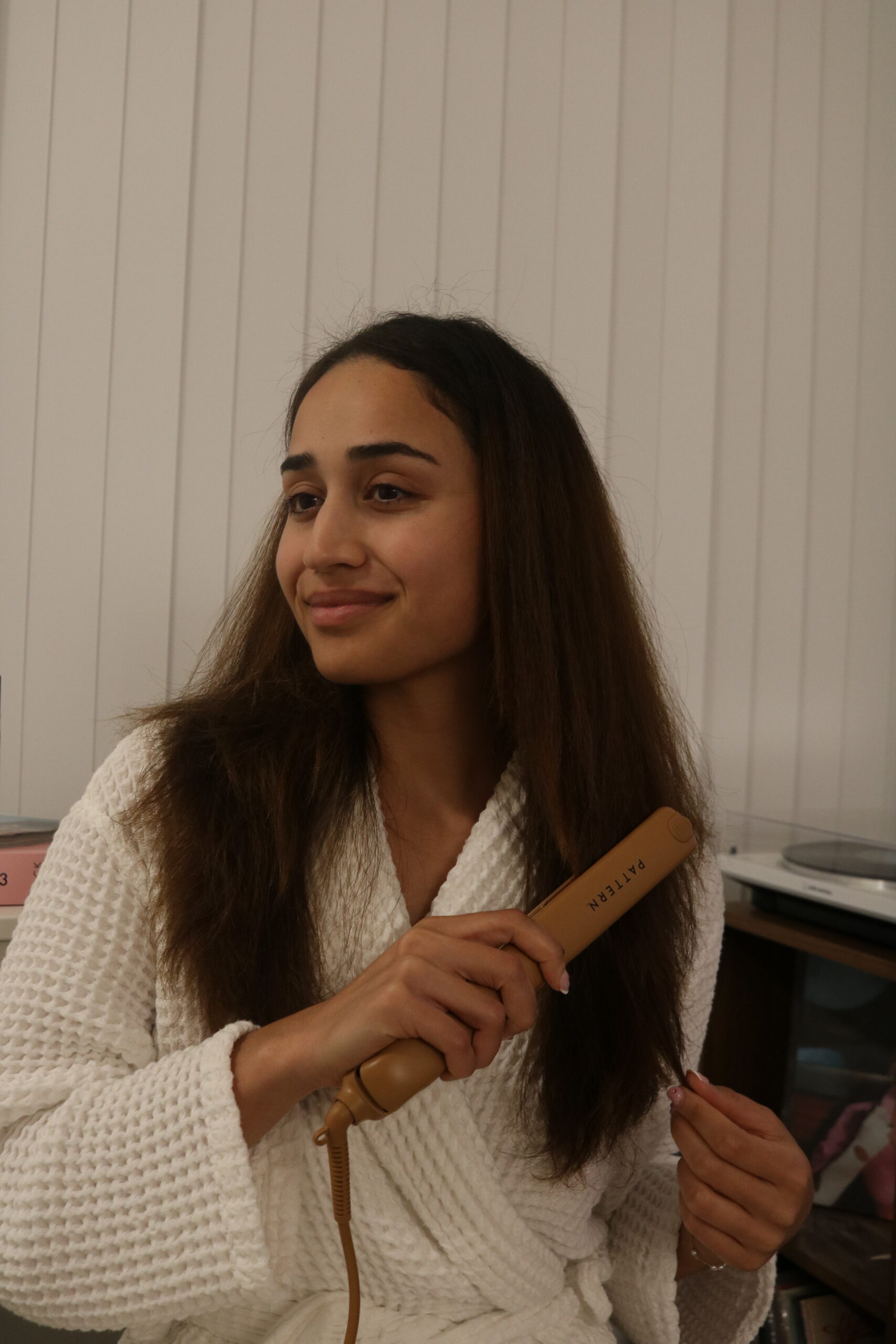 A woman in a white robe uses a flat iron to straighten her hair while sitting in a room with vertical blinds in the background.