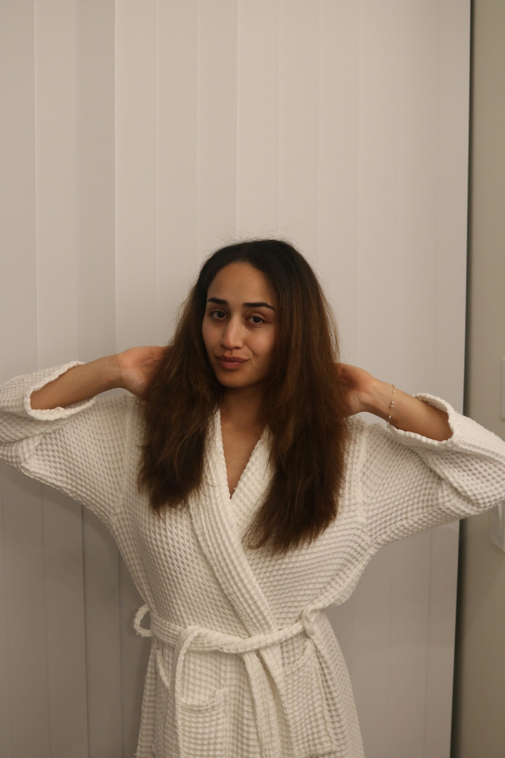 A person wearing a white bathrobe stands indoors, facing the camera with their hands raised to touch their hair, in front of vertical white blinds.