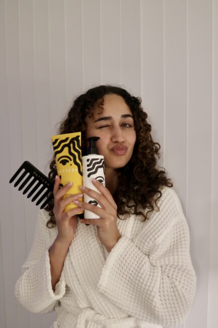 A person in a white robe holds a wide-tooth comb, a yellow tube, and a white bottle of hair products, posing in front of a white paneled background.