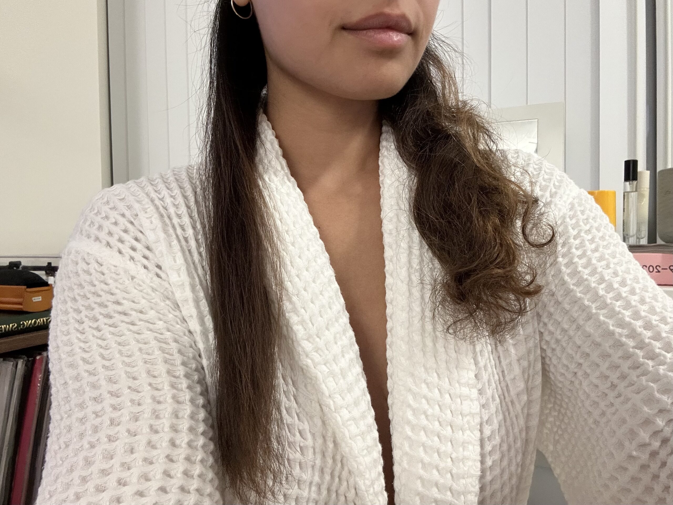 A person in a white robe shows their hair, with one half straight and the other half curly, against a background of shelves and books.