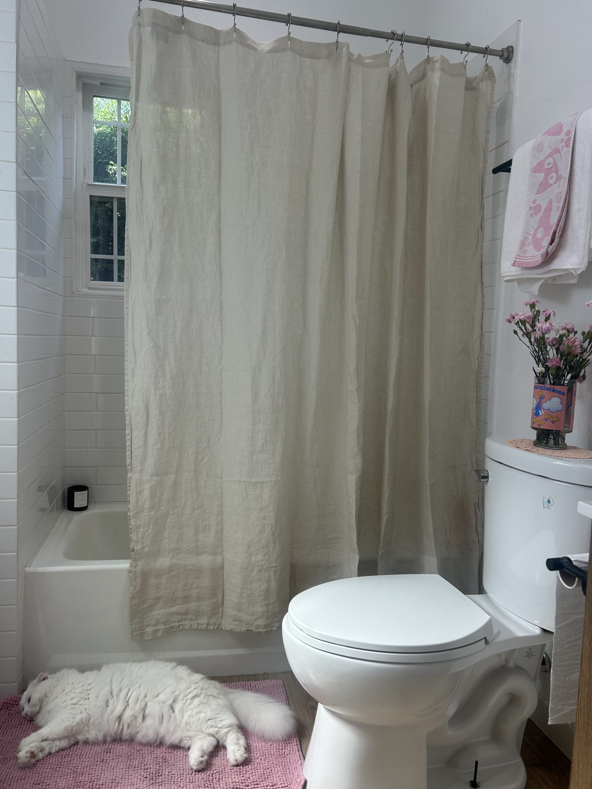 A white cat lies on a pink rug beside a toilet in a bathroom with a bathtub, beige shower curtain, pink towels, and flowers on the toilet tank.