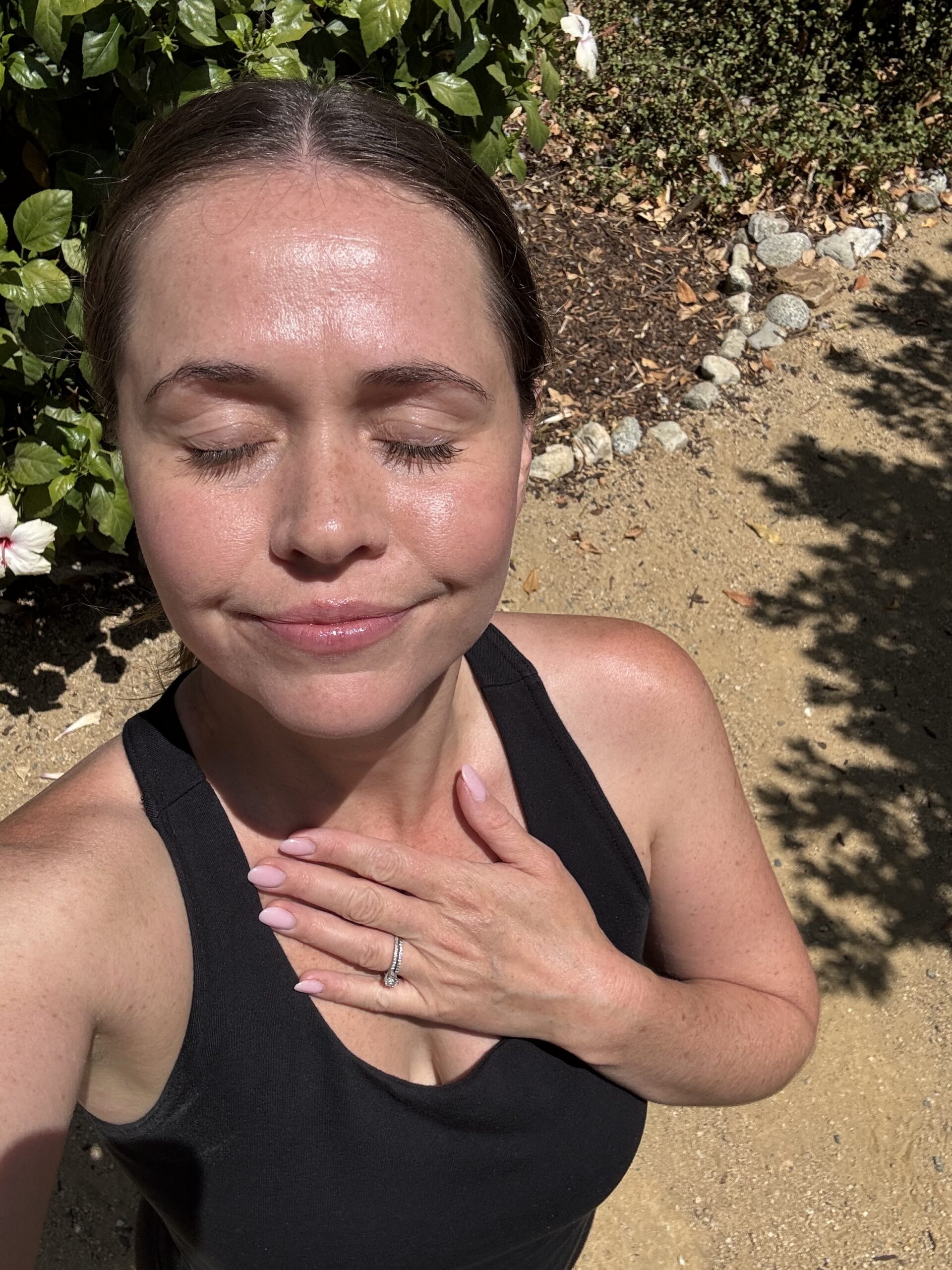 A woman in a black tank top stands outside with her eyes closed, hand on her chest, and sunlight on her face. Green leaves and a dirt path are visible in the background.