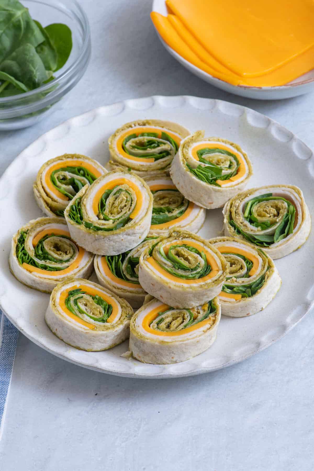 Plate of tortilla pinwheels filled with cheese and spinach, arranged in a circular pattern. Slices of cheese and a bowl of spinach are visible in the background.