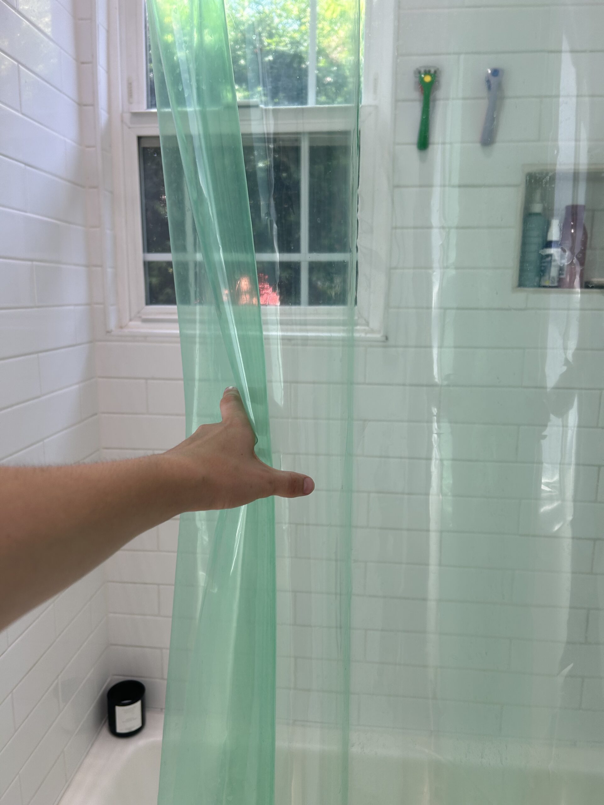 A hand pulls back a translucent green shower curtain in a white-tiled bathroom with a window and toiletries on a shelf.
