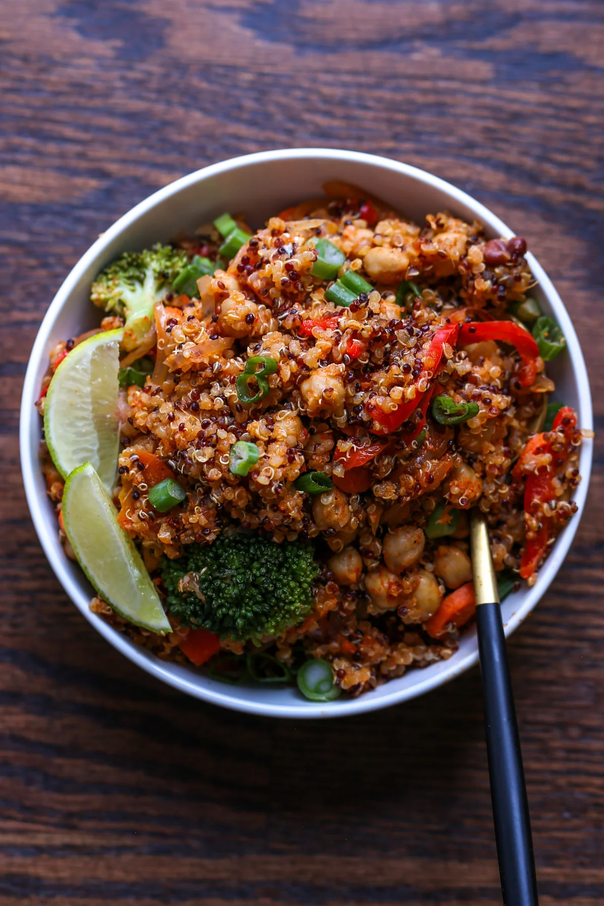 A bowl of quinoa stir-fry with broccoli, chickpeas, red bell peppers, green onions, and lime wedges on a wooden surface, with chopsticks resting on the side.