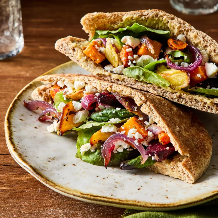 Two whole wheat pita halves filled with lettuce, roasted vegetables, red onions, and crumbled cheese are served on a ceramic plate on a wooden table.