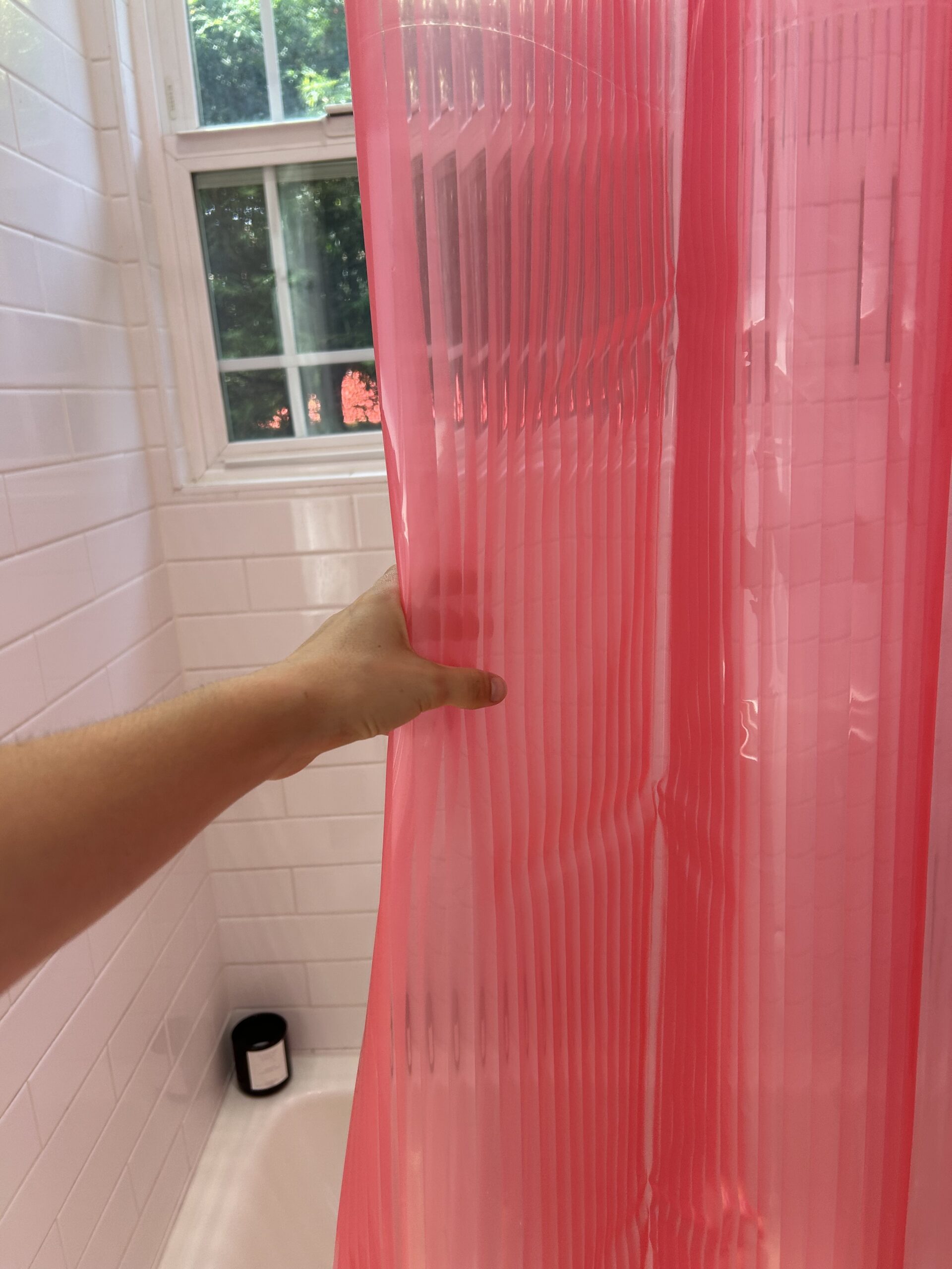 A hand pulls back a pink shower curtain in a white-tiled bathroom with a window and a black container on the tub’s edge.
