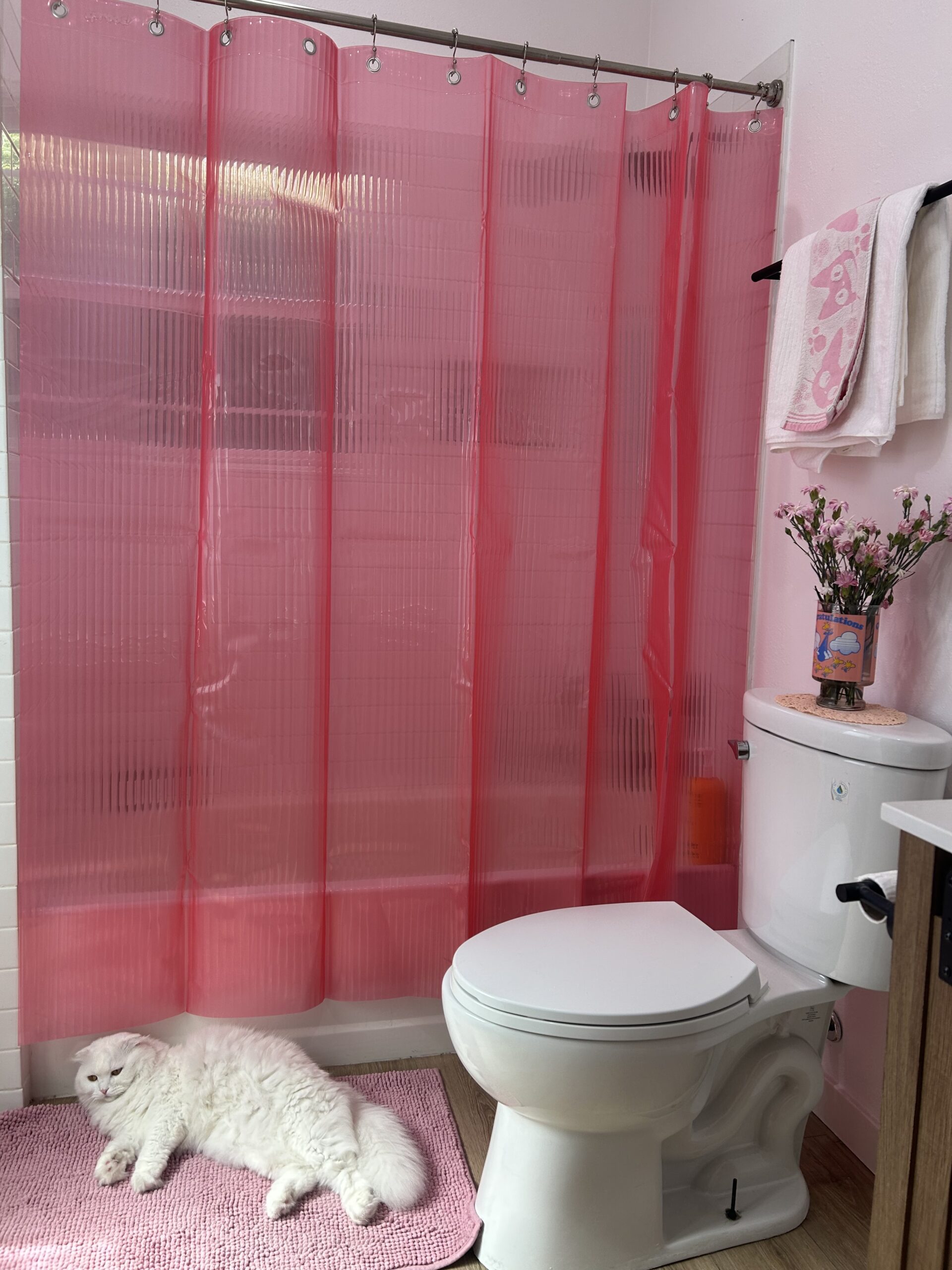 A fluffy white cat lies on a pink bath mat next to a toilet in a bathroom with a pink shower curtain, flowers, and folded towels.