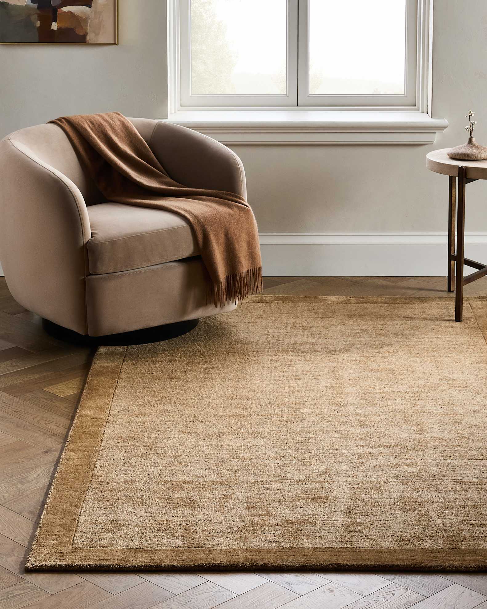 A beige armchair with a brown blanket draped over it sits on a tan area rug next to a round side table in a room with a window and parquet wood flooring.