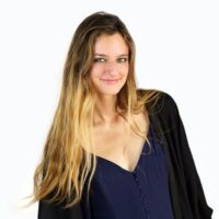 Young woman with long, blonde hair wearing a dark blue dress and black cardigan, standing against a plain white background.
