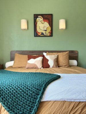 A neatly made bed with earth-toned pillows, a green knitted blanket, and a modern abstract painting on a green wall above the headboard. Two wall sconces flank the artwork.