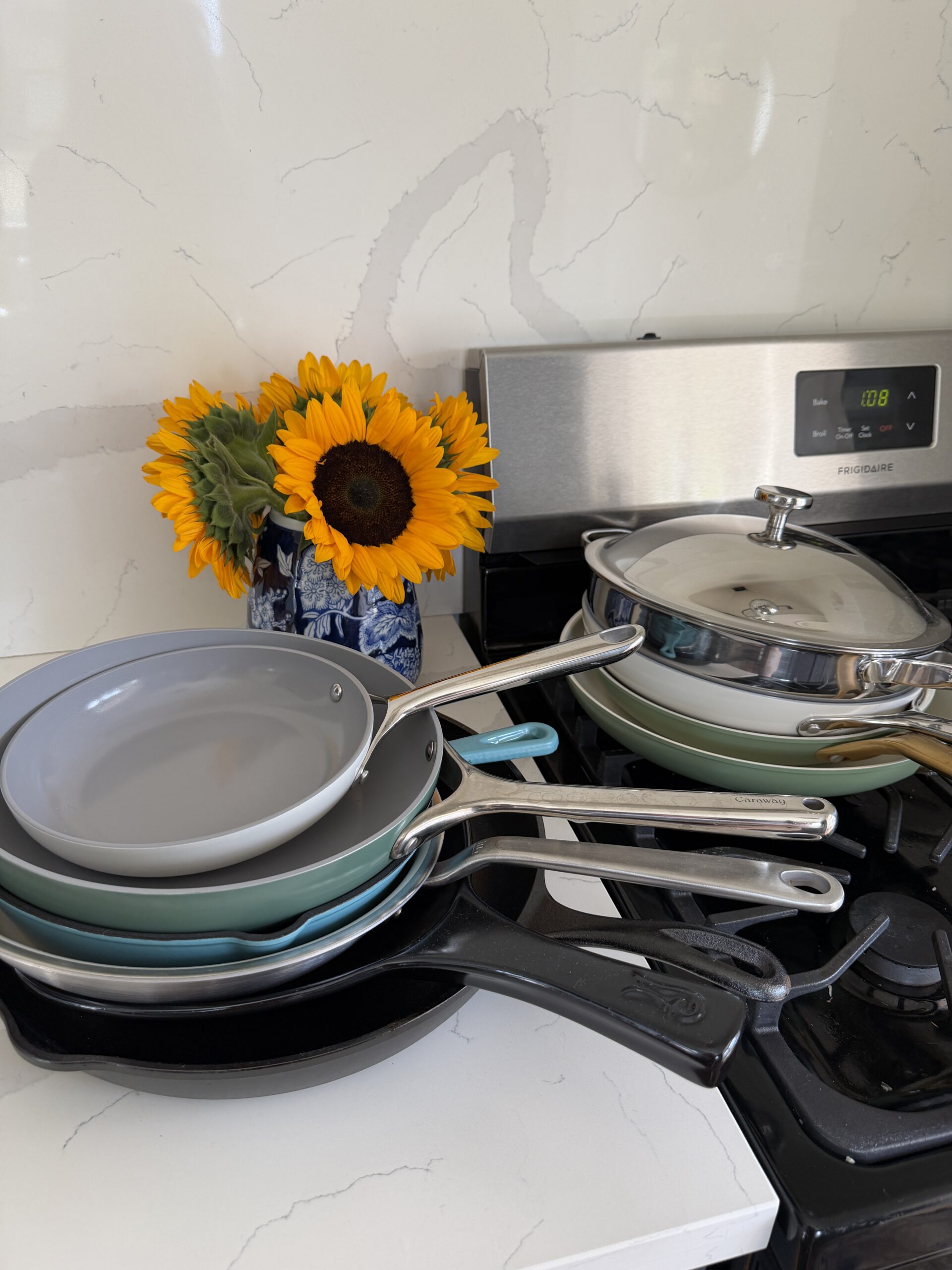 Four frying pans stacked on a stove next to a vase of sunflowers; two have gray nonstick surfaces, one has a lid, and one is a black cast iron skillet.