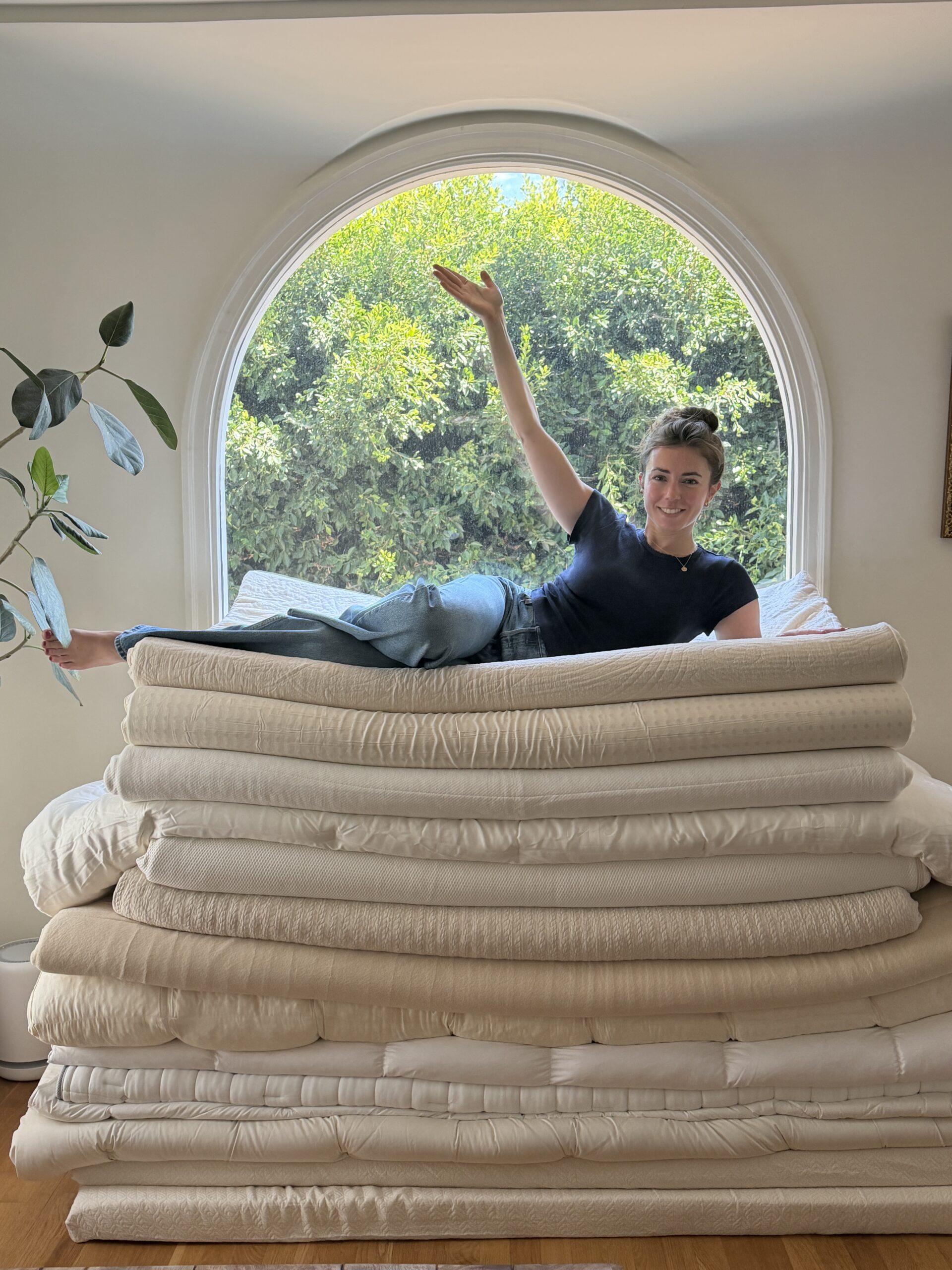 A person in a black shirt and jeans lies atop a tall stack of folded bedding in front of a large arched window with greenery outside.
