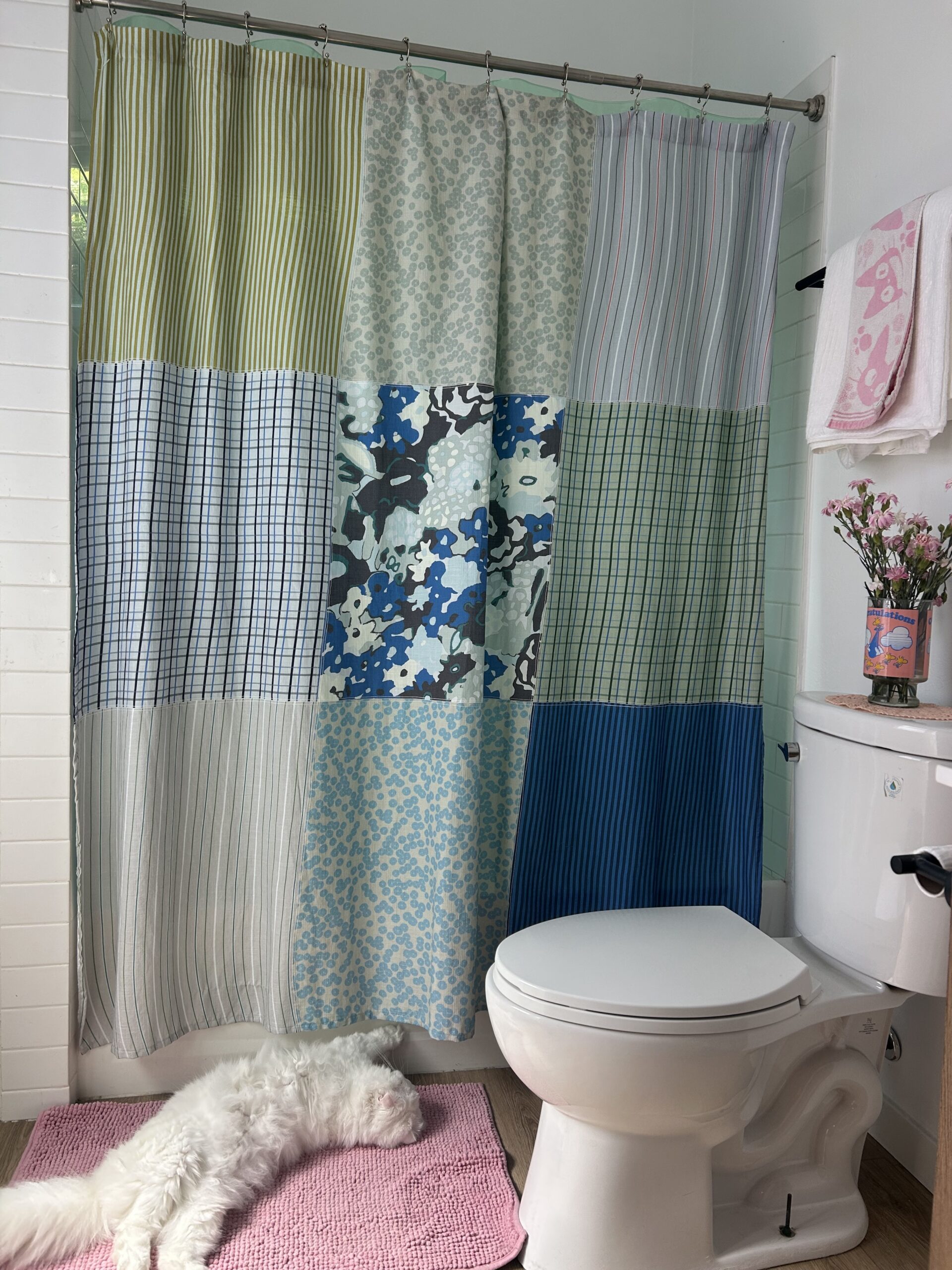 A white cat lies on a pink rug in a bathroom with a patchwork shower curtain, toilet, and towel hanging on the wall.
