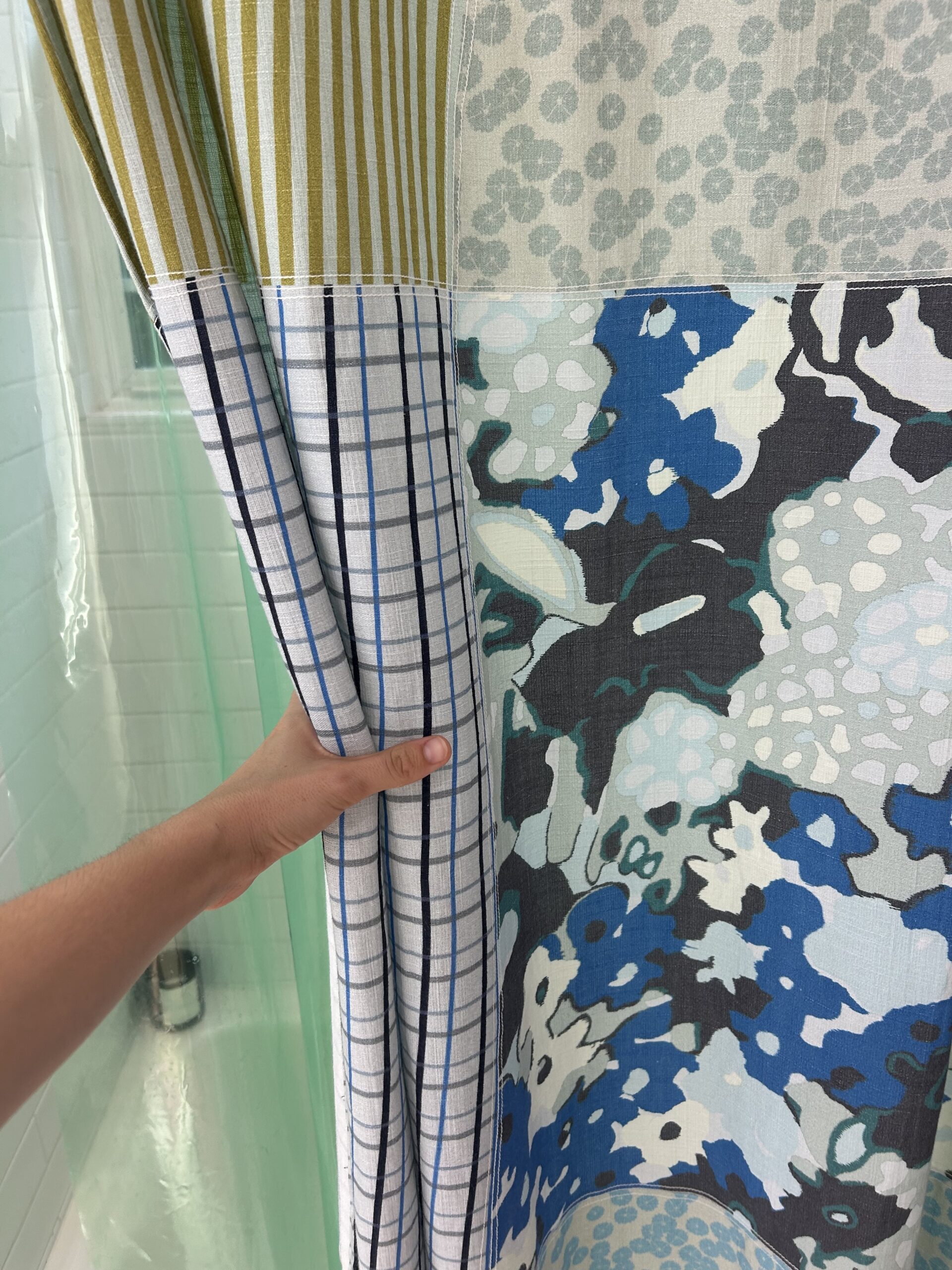 A hand pulls back a multicolored shower curtain with patchwork patterns, revealing a tiled wall and part of a shower.