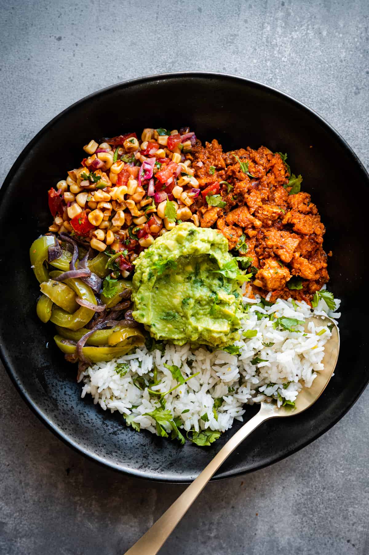 A black bowl with rice, sautéed green peppers and onions, seasoned ground meat, corn salsa, and guacamole, with a spoon on the side.
