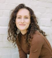 A woman with curly brown hair wearing a brown long-sleeve top and dark jeans sits in front of a white brick wall, smiling at the camera.