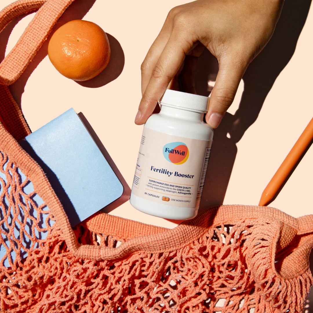A hand takes a FullWell Fertility Booster bottle from a coral net bag, with an orange, a blue notebook, and an orange pen nearby on a light surface.