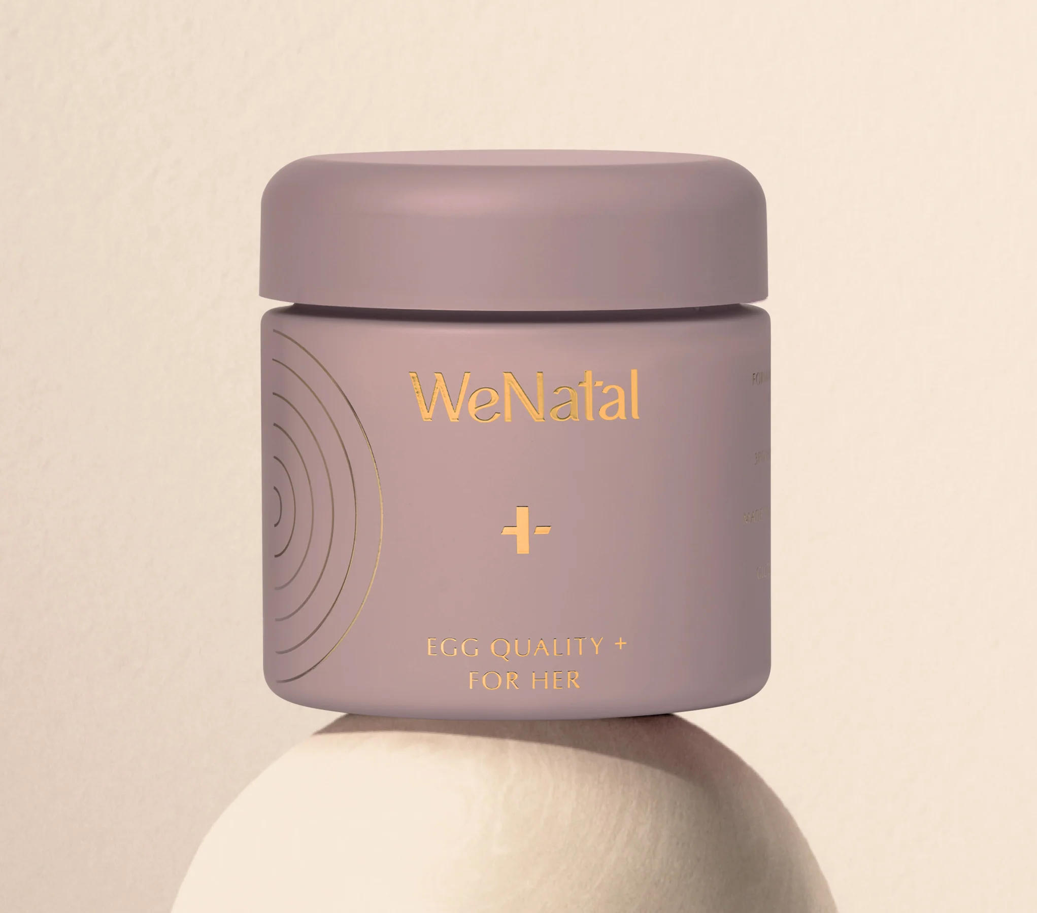 A mauve jar labeled “WeNatal Egg Quality + For Her” with gold text sits on a round white surface against a beige background.