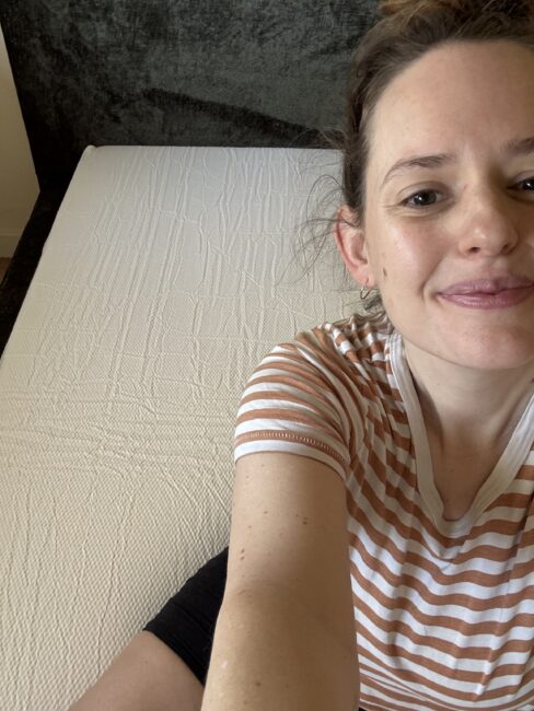 Person wearing a striped shirt sits on the edge of a bare mattress, smiling at the camera. The background includes a dark headboard.