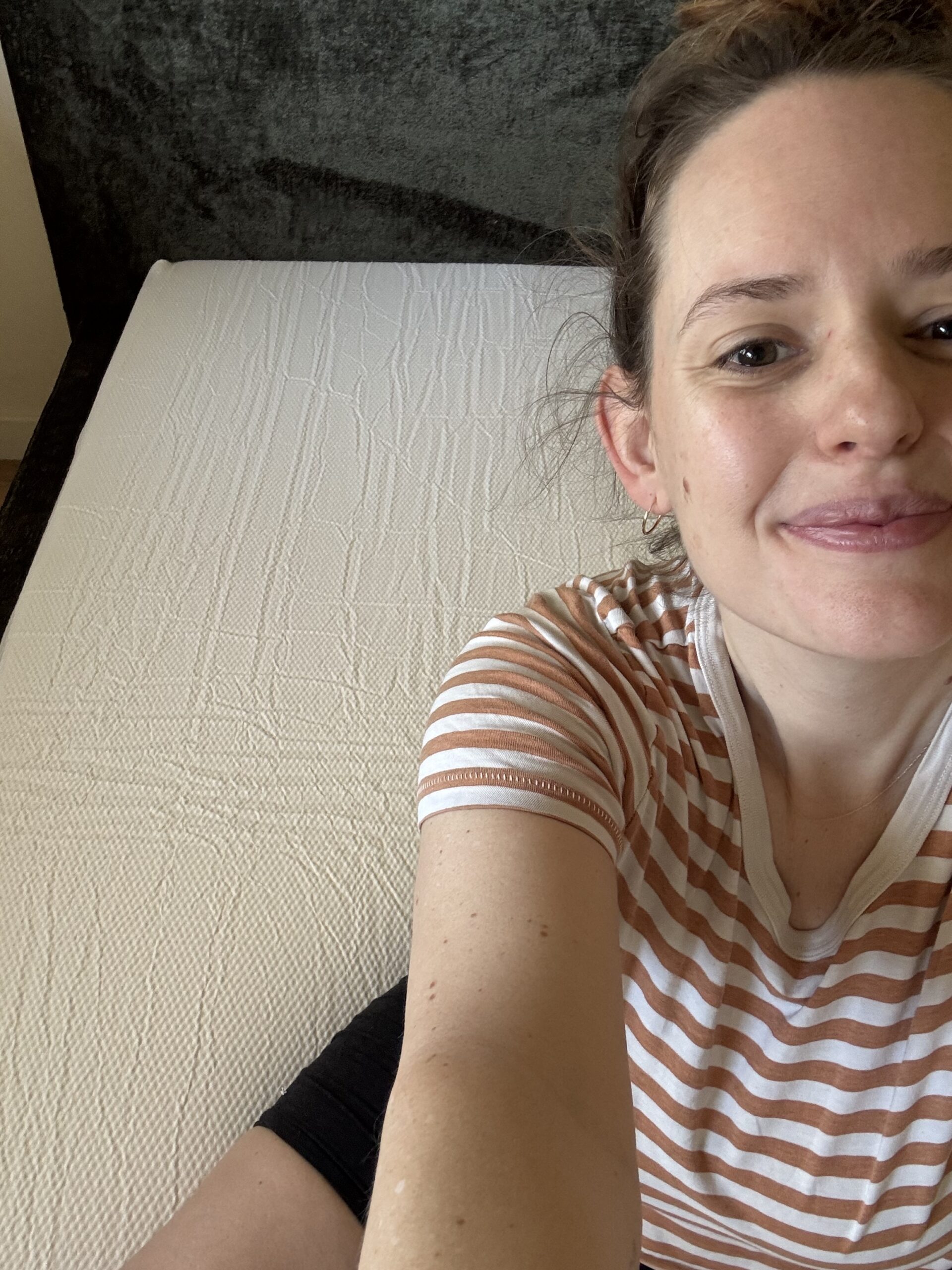 Person wearing a striped shirt sits on the edge of a bare mattress, smiling at the camera. The background includes a dark headboard.