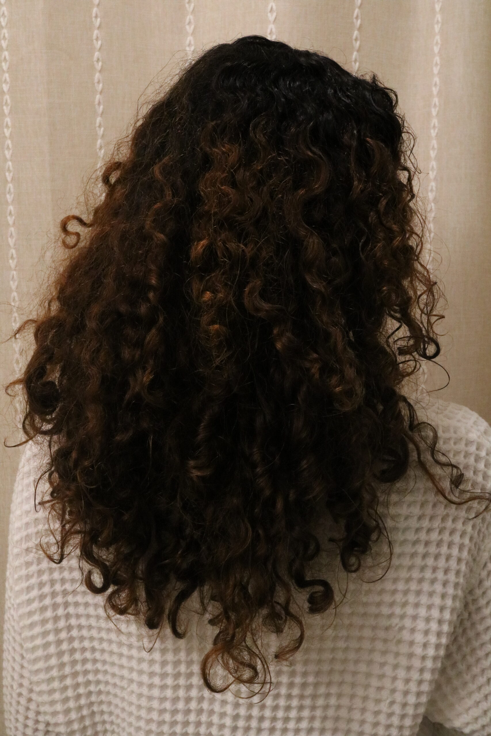 Person with curly dark brown hair, seen from behind, wearing a white textured sweater, standing in front of a beige curtain with a vertical pattern.