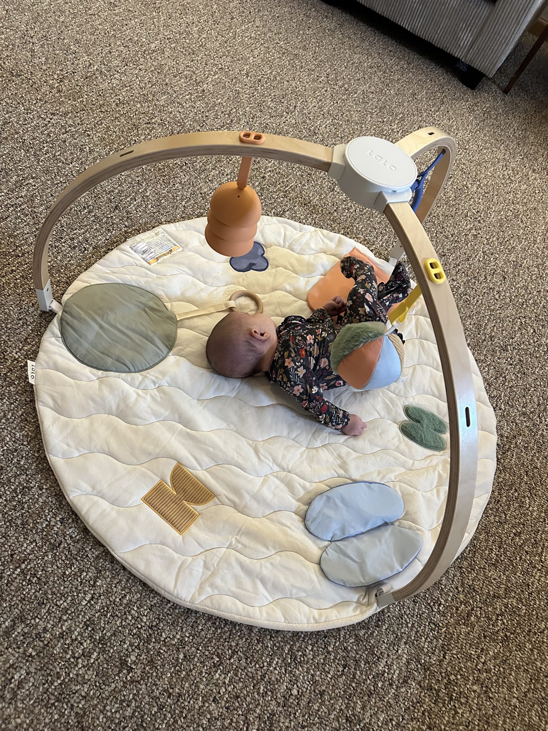 A baby lying on their stomach on a round play mat with an arched wooden activity gym.