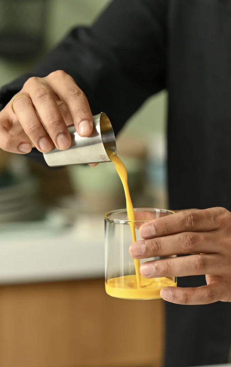 A person pours a thick yellow liquid from a small metal cup into a clear glass while holding both containers with their hands.