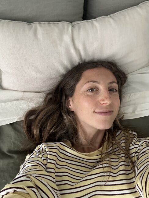 A woman with long brown hair and a striped shirt lies on a bed, looking at the camera, with white pillows visible behind her.
