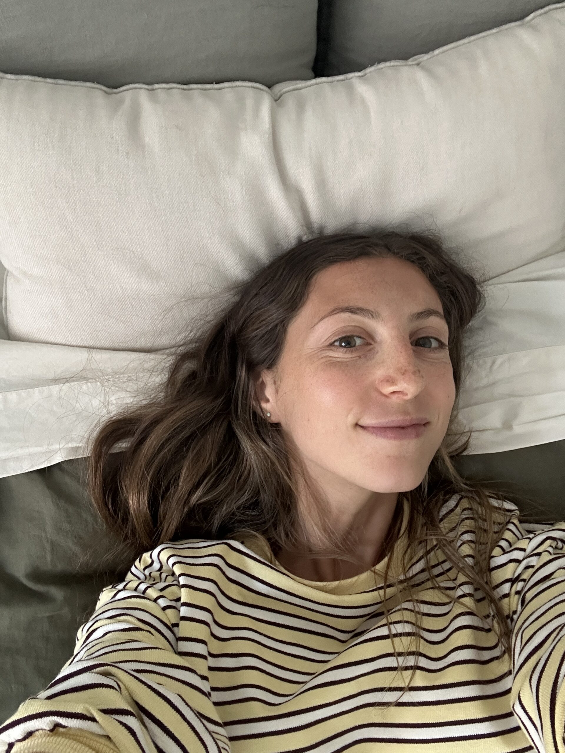 A woman with long brown hair and a striped shirt lies on a bed, looking at the camera, with white pillows visible behind her.