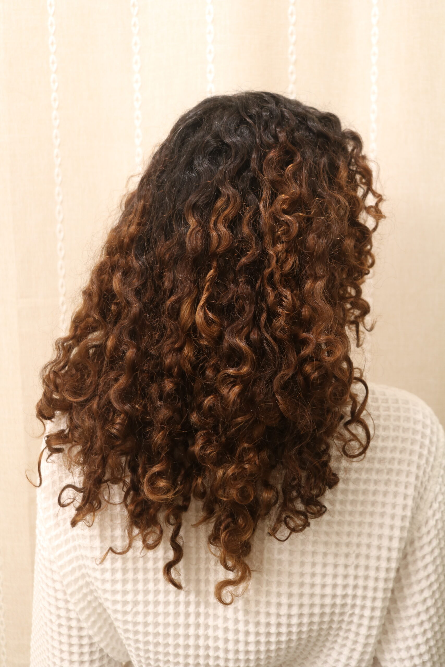 Person with shoulder-length, dark brown curly hair featuring lighter brown highlights, wearing a textured white top, shown from behind against a beige background.
