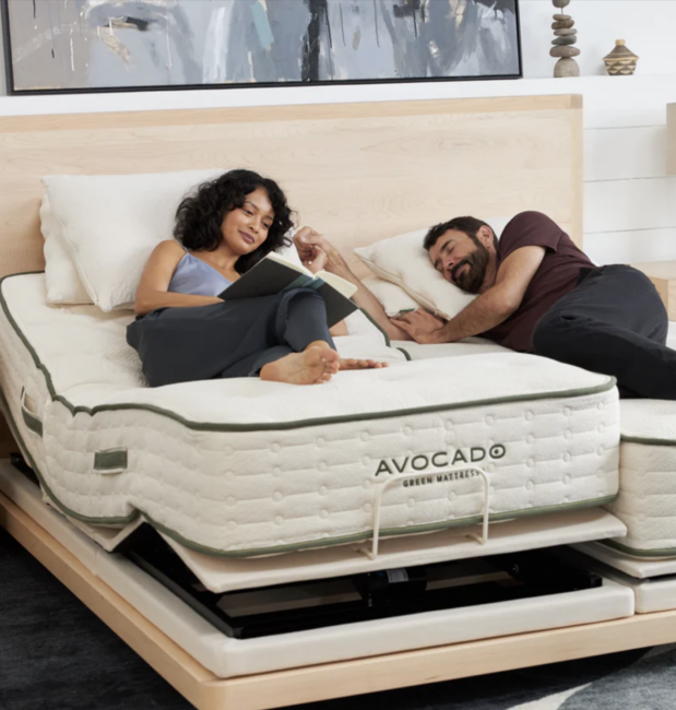 A woman reads on an adjustable Avocado mattress while a man sleeps beside her on a separate section of the split bed in a modern bedroom.