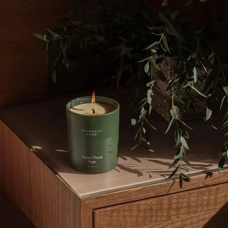 A green candle labeled “Sweet Floral Sage” burns on a wooden surface next to a plant with long, thin leaves.